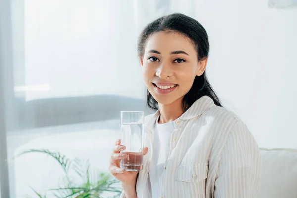 Позитивная латинская женщина держит стакан воды, улыбаясь в камеру — стоковое фото