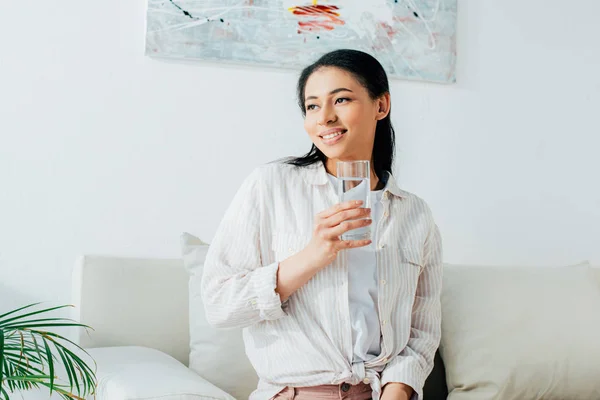 Belle femme de salon souriante et regardant loin tout en tenant un verre d'eau — Photo de stock