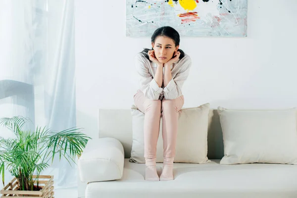 Sad latin woman sitting on back of sofa and looking away at home — Stock Photo