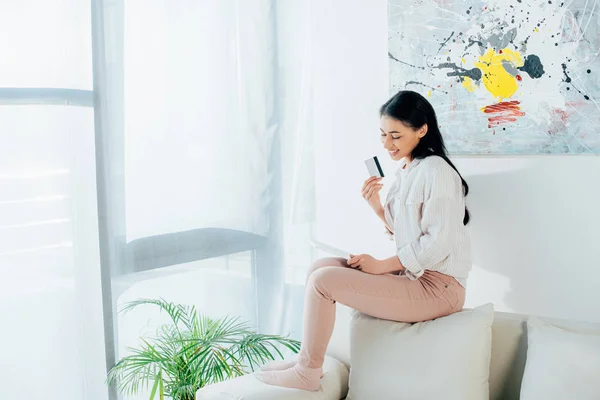 Felice donna latina in possesso di carta di credito mentre seduto sul retro del divano a casa — Foto stock