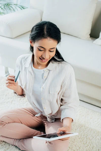 Femme latine souriante tenant une carte de crédit et utilisant une tablette numérique tout en étant assis sur le sol à la maison — Photo de stock