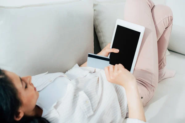 Mujer latina joven sosteniendo tarjeta de crédito y tableta digital con pantalla en blanco mientras está acostada en el sofá en casa - foto de stock