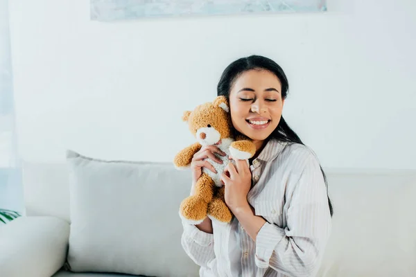 Feliz mujer latina con los ojos cerrados abrazando oso de peluche mientras está sentado en el sofá en casa - foto de stock