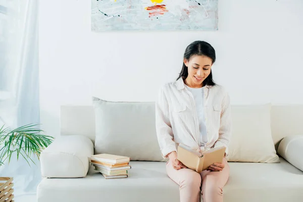 Bella donna latina seduta sul divano a casa, sorridente e libro di lettura — Foto stock
