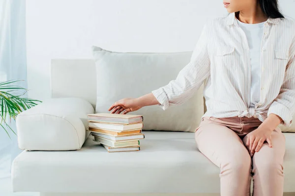 Abgeschnittene Aufnahme einer jungen lateinamerikanischen Frau, die auf einem Sofa in der Nähe eines Bücherstapels sitzt — Stockfoto