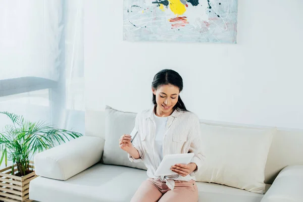 Mujer latina sonriente sosteniendo tarjeta de crédito y usando tableta digital mientras está sentada en el sofá en casa - foto de stock