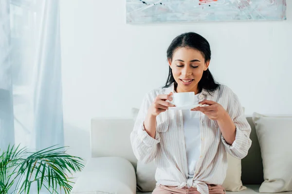 Bonita mujer latina bebiendo café con los ojos cerrados mientras está sentada en el sofá en casa - foto de stock