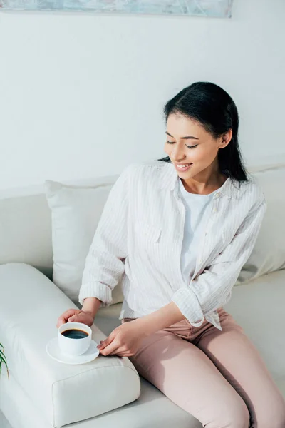 Cheerful latin woman holding cup of coffee while sitting on couch at home — Stock Photo