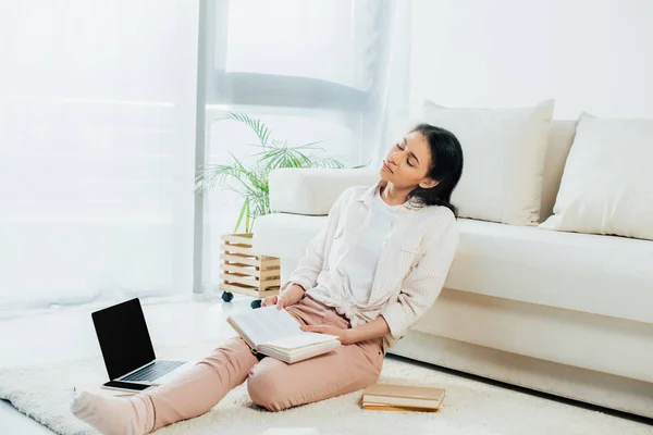 Mujer latina cansada sosteniendo libro abierto mientras está sentado en el suelo cerca de la computadora portátil — Stock Photo