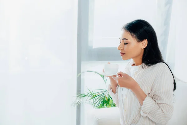 Pretty latin woman holding coffee cup with closed eyes while standing by window at home — Stock Photo
