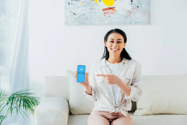 KYIV, UKRAINE - APRIL 26, 2019: Attractive latin woman pointing with finger at smartphone with Skype app on screen. — Stock Photo