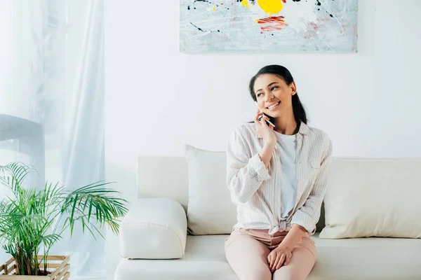 Hermosa mujer latina hablando en el teléfono inteligente mientras está sentada en el sofá y mirando hacia otro lado - foto de stock