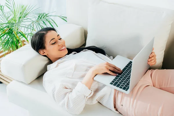 Mujer latina feliz usando el ordenador portátil mientras está acostado en el sofá en casa - foto de stock