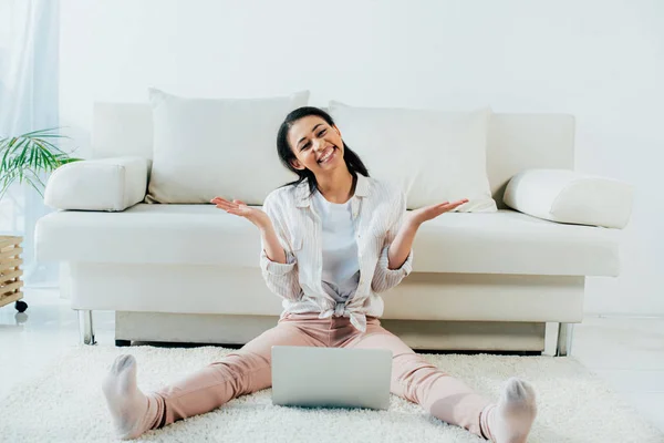 Happy latin woman gesturing and smiling at camera while sitting on floor and using laptop — Stock Photo
