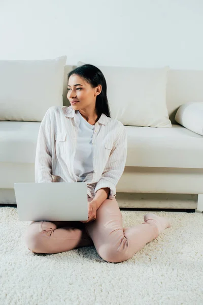 Sorrindo mulher latina olhando para longe enquanto sentado no chão e usando laptop — Fotografia de Stock