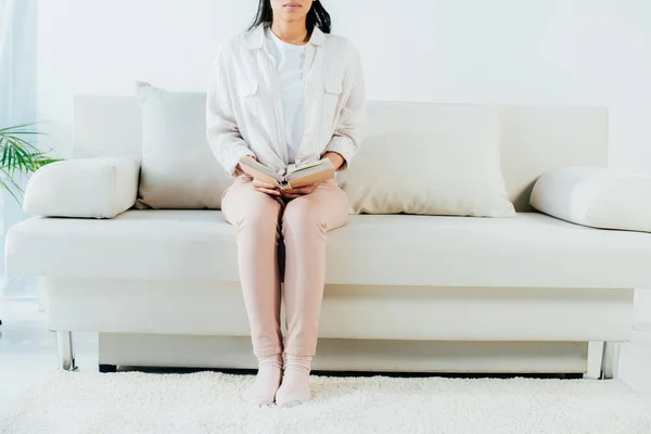 Cropped view of young woman holding book while sitting on sofa at home — Stock Photo