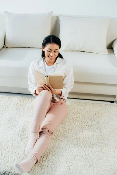 Attrayant latin femme assise lecture livre et sourire tout en étant assis sur le sol à la maison — Photo de stock