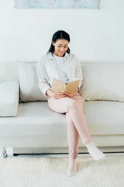 Pretty latin woman reading book while sitting on sofa at home — Stock Photo