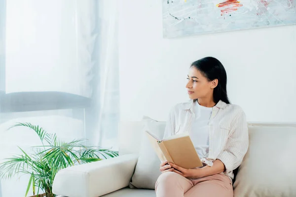 Young latin woman looking away and smiling while sitting on sofa and holding book — Stock Photo