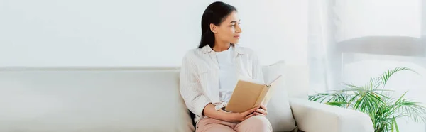 Plano panorámico de la mujer latina bonita sosteniendo libro mientras está sentado en el sofá y mirando hacia otro lado - foto de stock