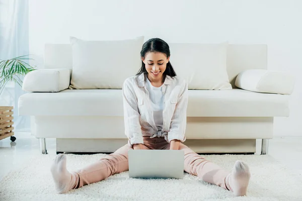 Mujer latina alegre usando el ordenador portátil mientras está sentado en el suelo en casa - foto de stock