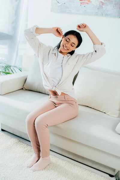 Bonita mujer latina sosteniendo las manos levantadas y sonriendo mientras está sentada en el sofá en casa - foto de stock