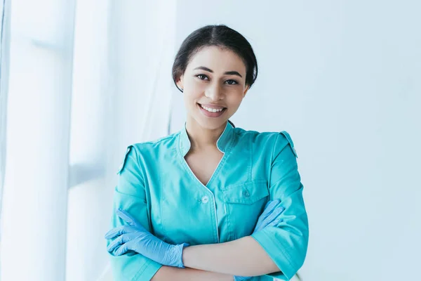 Positive latin doctor in latex gloves standing with crossed arms and smiling at camera — Stock Photo