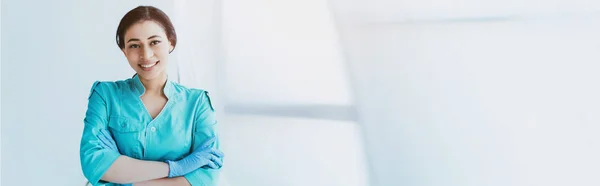 Plano panorámico de alegre médico latino de pie con los brazos cruzados y sonriendo a la cámara - foto de stock