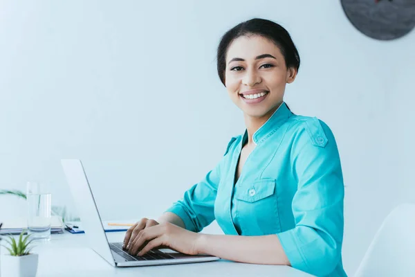 Attraente medico latino utilizzando computer portatile sul posto di lavoro e sorridente alla fotocamera — Foto stock