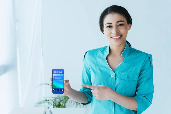 Sorrindo médico latino apontando com o dedo para o smartphone com aplicativo de reserva na tela — Fotografia de Stock