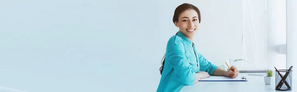 Plano panorámico del joven médico latino sonriendo a la cámara mientras está sentado en el lugar de trabajo - foto de stock