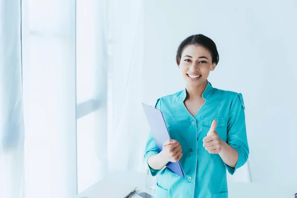 Happy latin doctor showing thumb up and smiling at camera while standing by window in hospital — Stock Photo