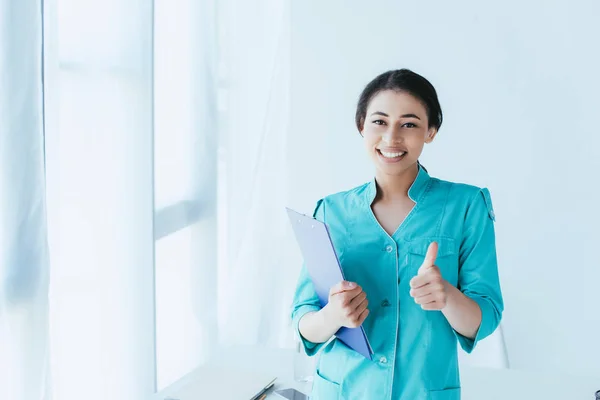 Feliz médico latino mostrando el pulgar hacia arriba y sonriendo a la cámara mientras está de pie junto a la ventana en el hospital - foto de stock