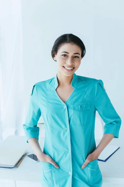 Hermosa doctora latina cogida de la mano en los bolsillos y sonriendo a la cámara - foto de stock