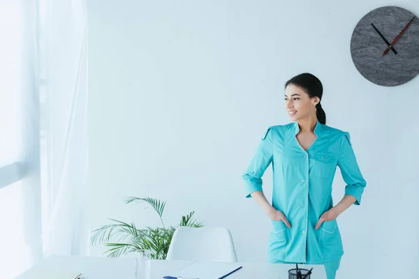 Cheerful latin doctor holding hands in pockets while standing by workplace and looking away — Stock Photo