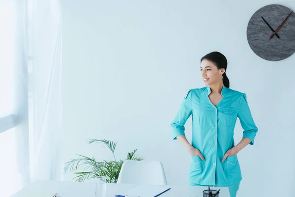 Alegre médico latino cogido de la mano en los bolsillos mientras está parado en el lugar de trabajo y mirando hacia otro lado - foto de stock