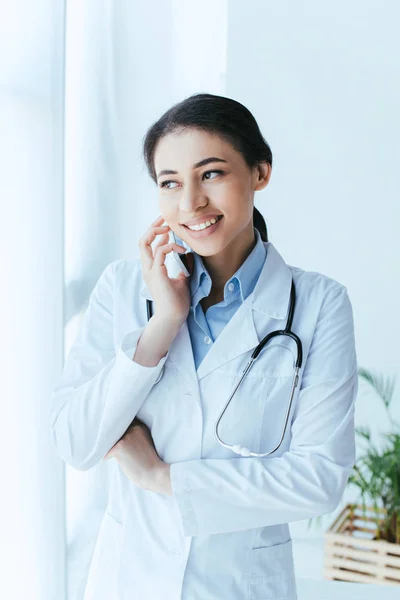 Médico latino positivo hablando en smartphone y mirando hacia otro lado mientras está de pie junto a la ventana en el hospital - foto de stock