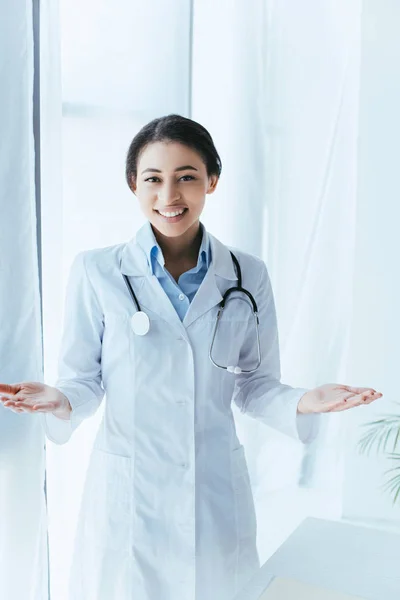 Alegre médico latino con estetoscopio mostrando gesto de bienvenida mientras sonríe a la cámara - foto de stock