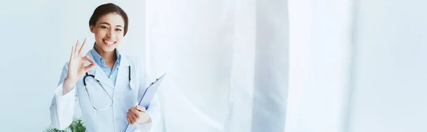 Tiro panorámico de médico latino feliz mostrando gesto bien y sujetando el portapapeles - foto de stock