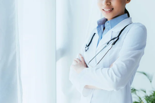 Обрезанный снимок молодого латинского врача со стетоскопом, стоящего у окна со скрещенными руками — стоковое фото