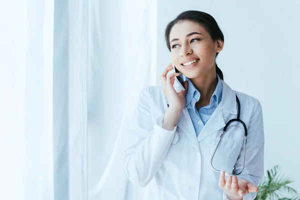 Hübsche junge Ärztin spricht auf Smartphone, während sie im Krankenhaus am Fenster steht — Stockfoto