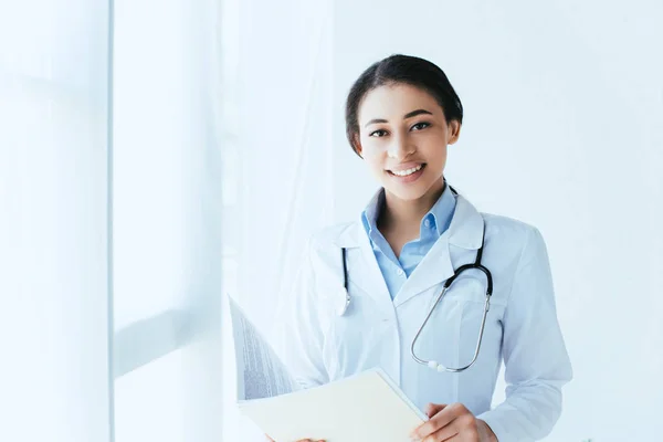 Alegre médico latino con estetoscopio sosteniendo la carpeta de papel y sonriendo a la cámara - foto de stock