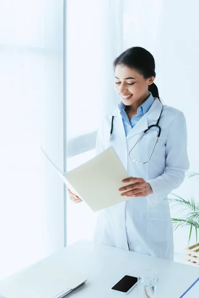 Sonriente médico latino sosteniendo la carpeta de papel mientras está de pie junto a la ventana en el hospital - foto de stock