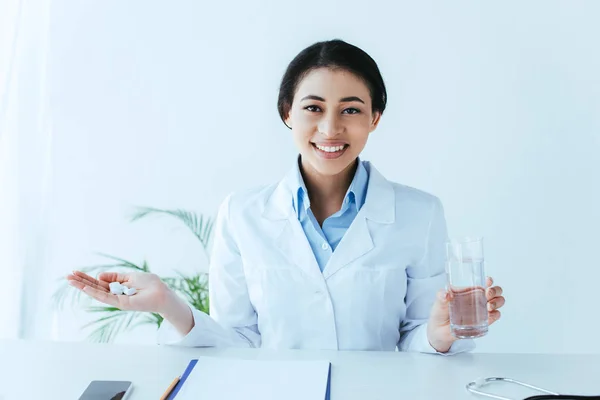 Alegre médico latino con un puñado de pastillas y un vaso de agua sentado en el lugar de trabajo y sonriendo a la cámara - foto de stock