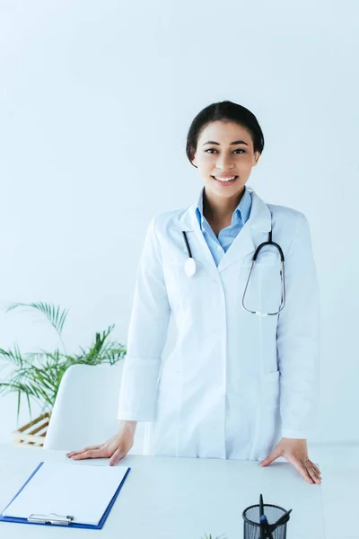Médico latino alegre com estetoscópio sorrindo para a câmera enquanto estava perto do local de trabalho no hospital — Fotografia de Stock