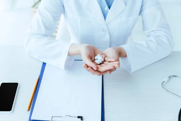 Vista parcial del médico con un puñado de pastillas sentadas en el lugar de trabajo cerca del portapapeles - foto de stock