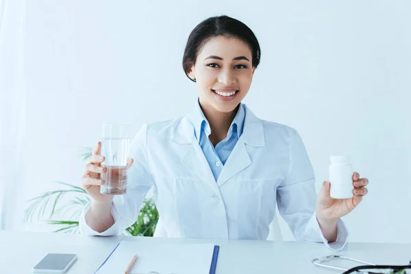 Alegre médico latino sosteniendo pastillas contenedor y vaso de agua mientras está sentado en el lugar de trabajo y sonriendo a la cámara - foto de stock