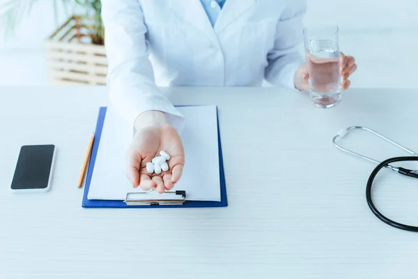 Plan recadré de jeune médecin avec un verre d'eau et une poignée de pilules assis sur le lieu de travail à l'hôpital — Photo de stock