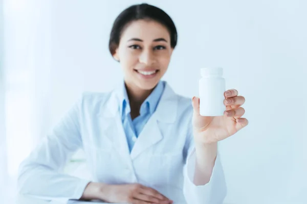 Enfoque selectivo de alegre médico latino sosteniendo pastillas contenedor y sonriendo a la cámara - foto de stock