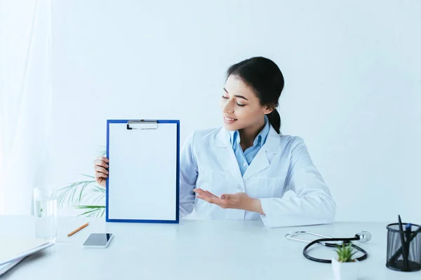Sonriente médico latino sujetando el portapapeles con papel en blanco mientras está sentado en el escritorio - foto de stock